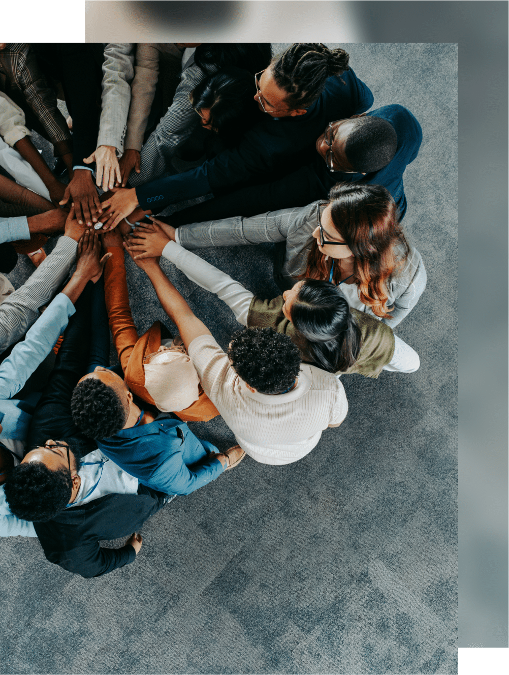 A diverse group of people places their hands together in a circle, symbolizing teamwork and unity, on a gray carpeted floor.