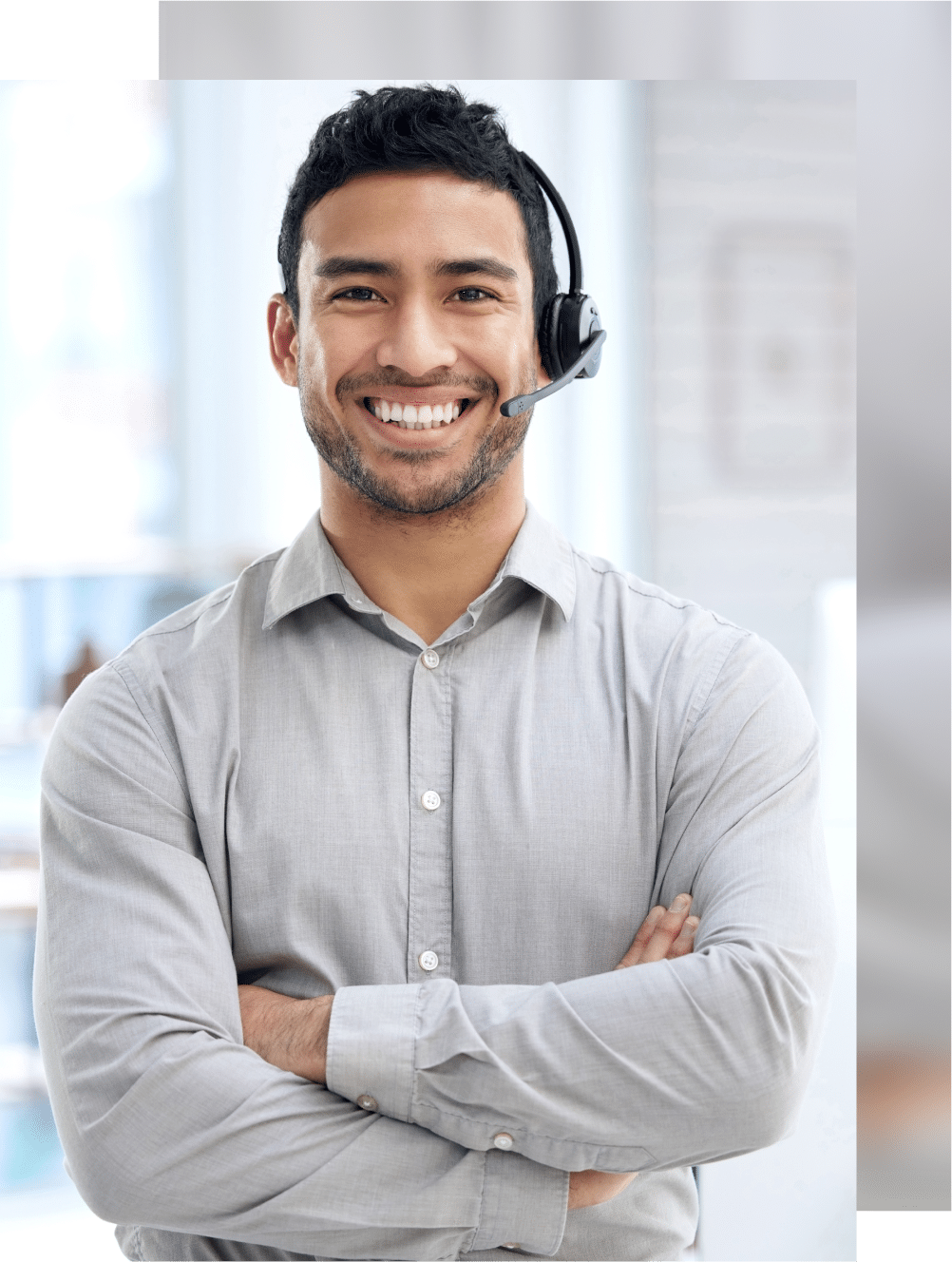 A smiling person wearing a headset and a light gray shirt stands with arms crossed in an office environment.