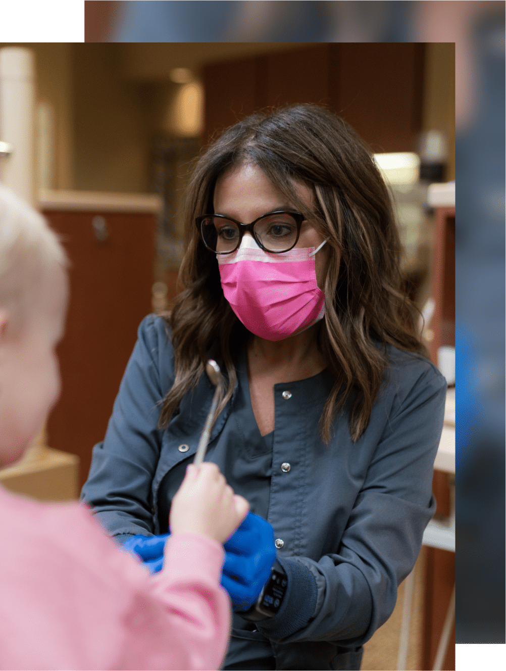 A person wearing a pink mask and glasses interacts with a child, holding a dental instrument in a clinic setting.